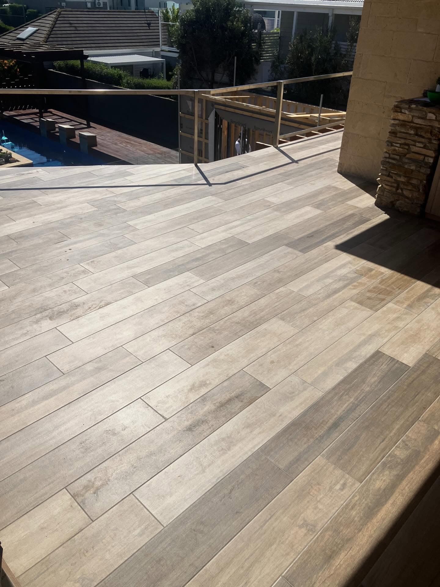 Timber-look tiles laid as a deck beside a pool, with a wire balustrade and rooflines behind