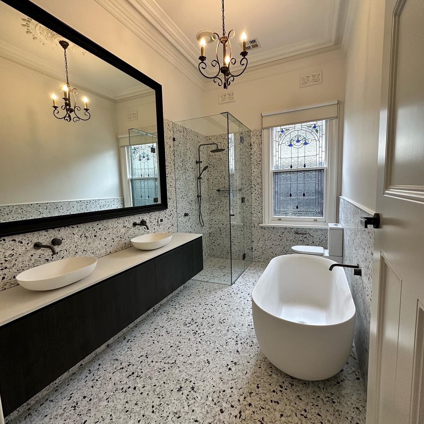 Bathroom with speckled terrazzo on the floor and walls, twin round basins on a black vanity, a full-width mirror and a freestanding bath