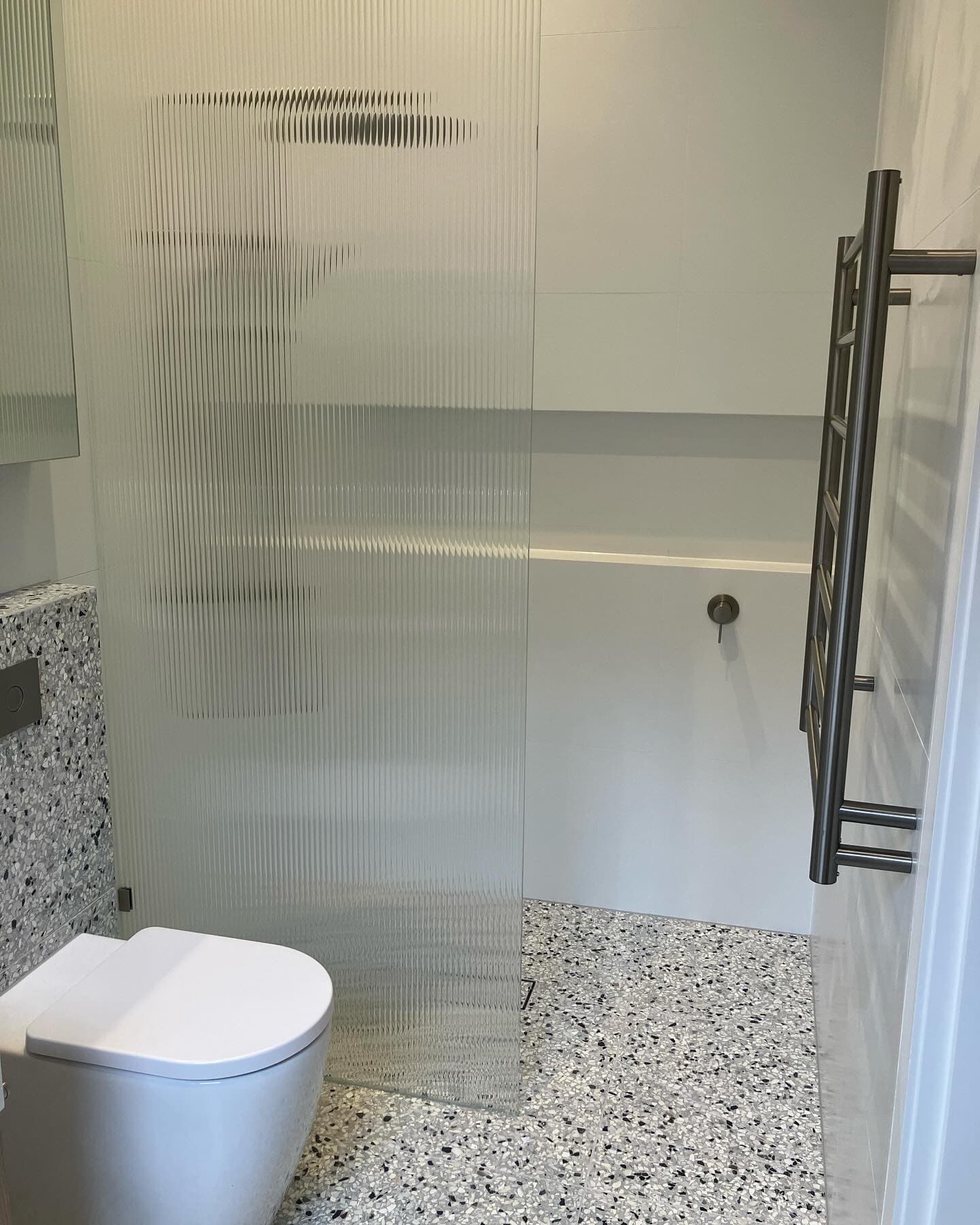 Walk-in shower behind a fluted glass screen, with a speckled terrazzo floor and a heated towel rail